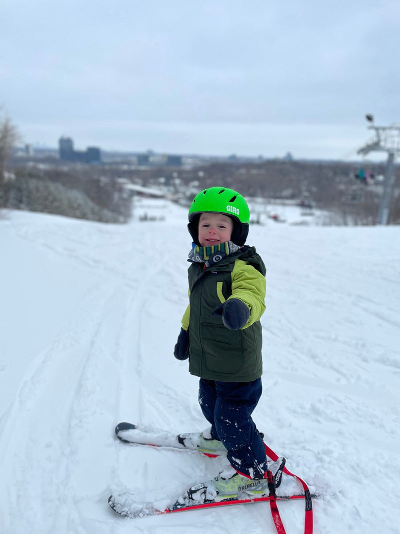 Toddler at the top of a ski hill wearing Snap Two Ski ankle tethers, ready for a first ski run
