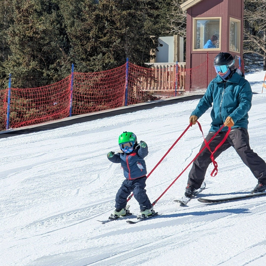 Parent and child skiing together with the Snap Two Ski ankle tether on a snow-covered slope with trees having fun on a sunny day.
